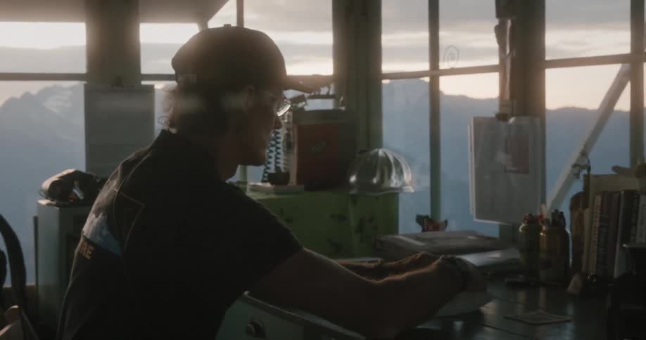 A man is sitting at a desk doing paperwork in a mountain watchtower at dusk