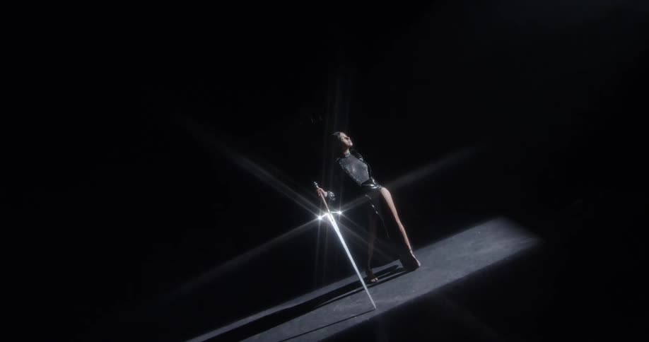 A woman poses with a large sword in a patch of a light in a dark room