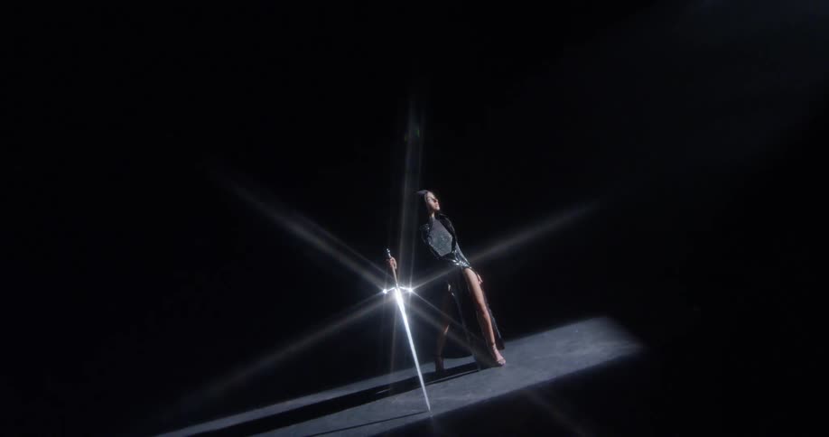 Woman stands on dark stage holding shining long sword lit by ceiling spotlight