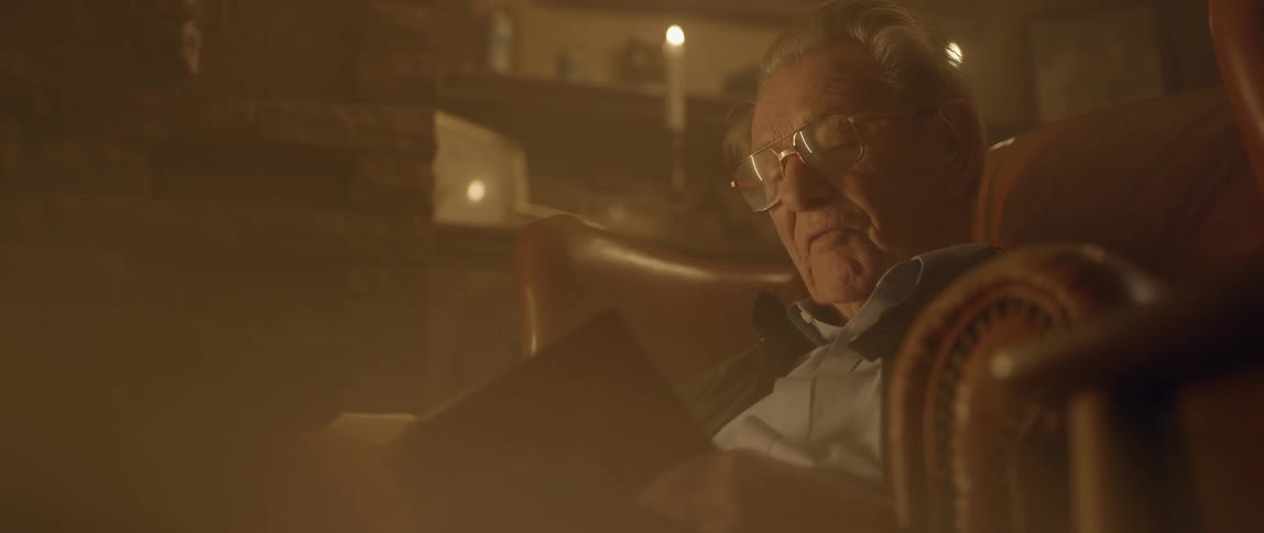 An elderly man reads a book out loud in a candle lit room