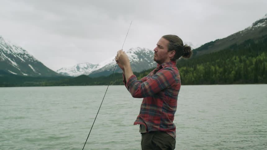 A man prepares his fishing rod as he stands next to the water in front of a mountain range