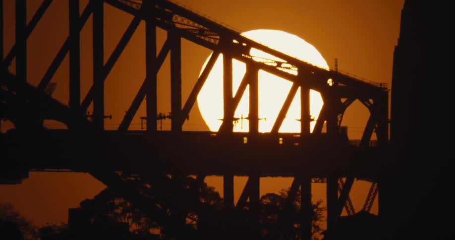 Silhouette of a train crossing a bridge with other traffic and a bright sun in an orange sky