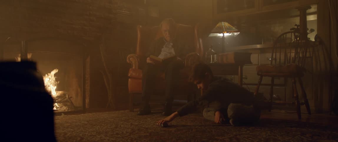 A child plays with a toy car in front of a burning fireplace with an elderly person sitting in the armchair behind him