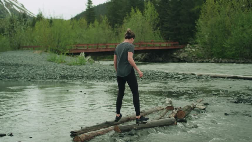 Woman with gray t shirt and black leggings and trainers walks on tree trunks on the river