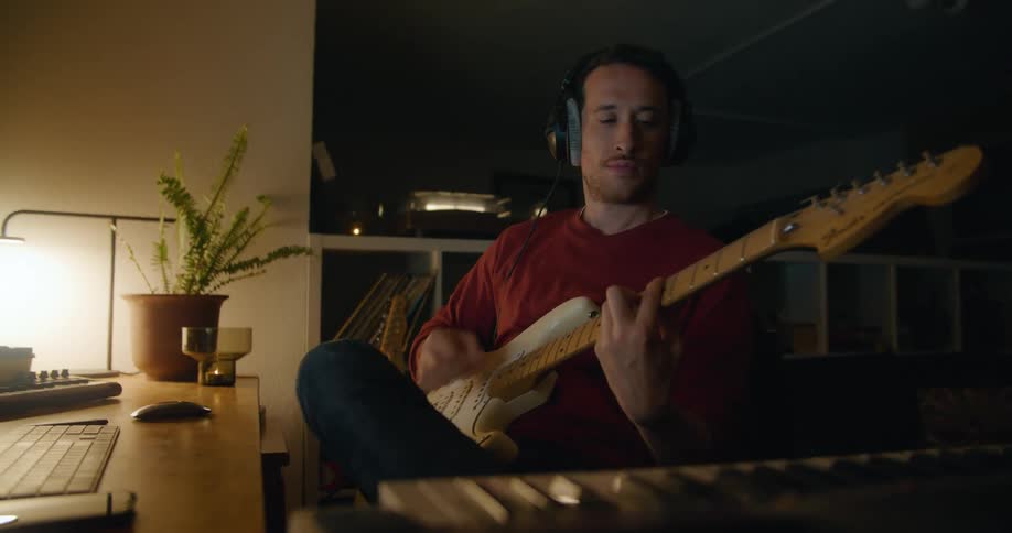 A man strums a guitar as he uses a computer on the desk