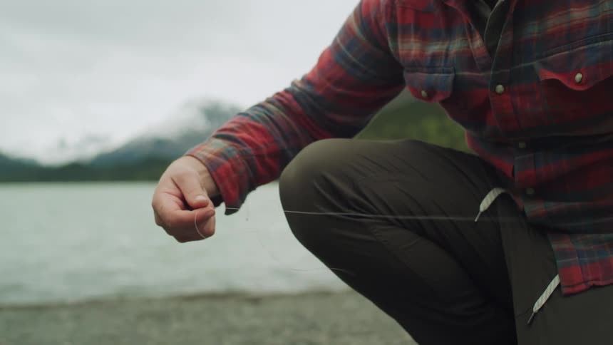 Crouching man untangles a fishing line near a lake in the mountains