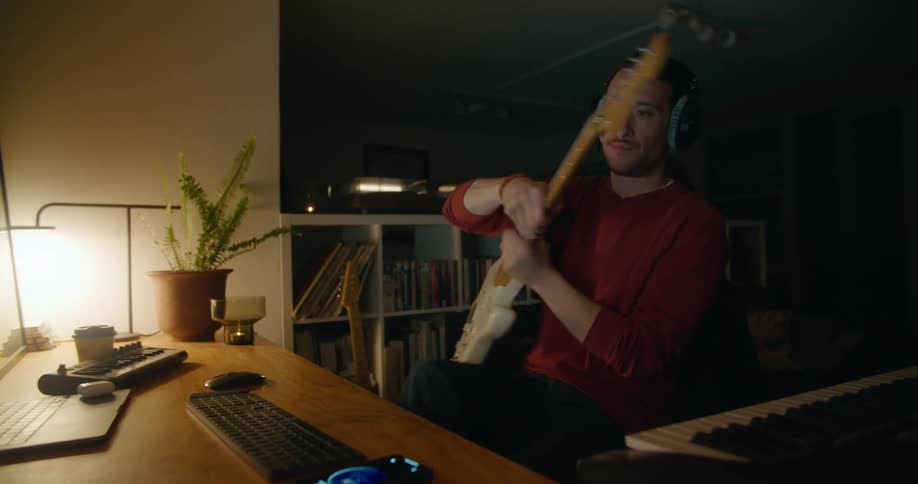 Man with red sweater sitting down and wearing headphones strums bass guitar looking at desktop in studio