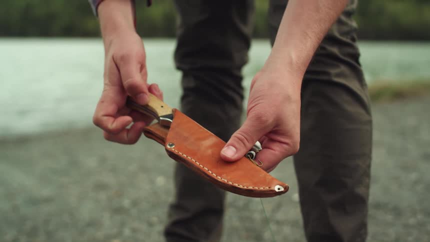 A man standing next to a lake carefully places a small knife back in its sheath