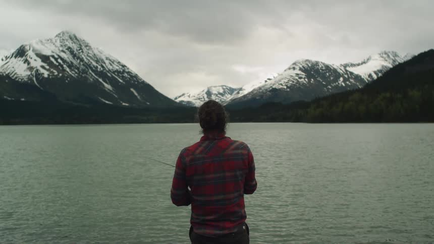A man fishes in a lake in a front of a mountain range