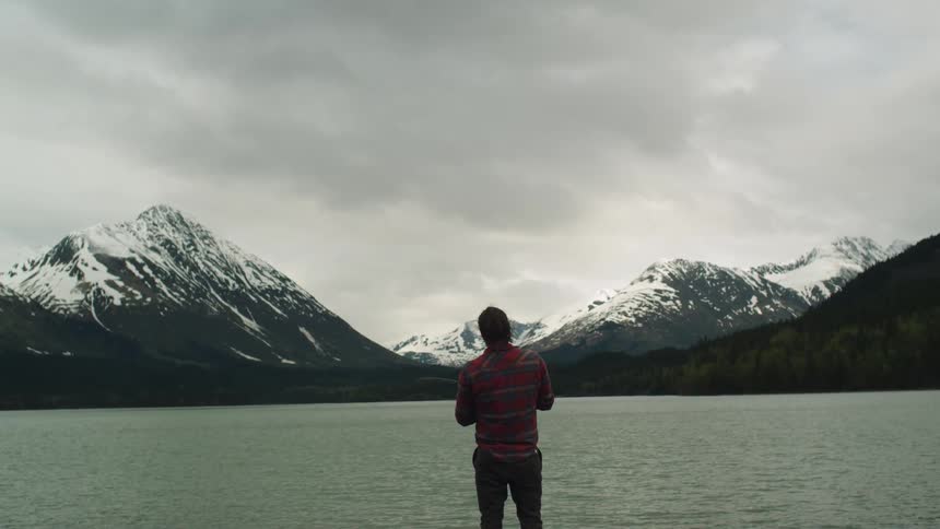 A man is standing looking at a lake near a snowy mountain range