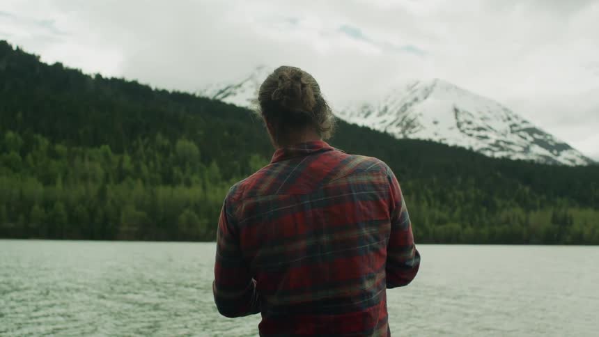 Man stands on shore fishing across river from forest and mountains