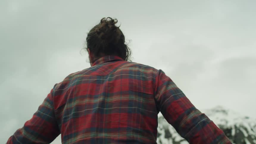 A man in a flannel shirt walks near a forested area and a group of snow capped mountains