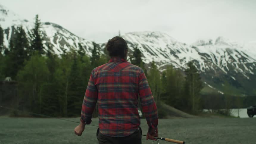 A man carrying a fishing rod walks toward a mountain lake