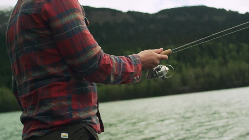 Man with plaid shirt and brown pants casts a fishing line into lake next to mountain with forest