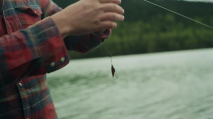 Man with plaid shirt puts fishing lure on fishing line next to river
