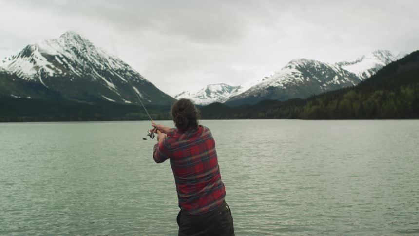 A man casts his fishing line into a lake with snow capped mountains on a cloudy day