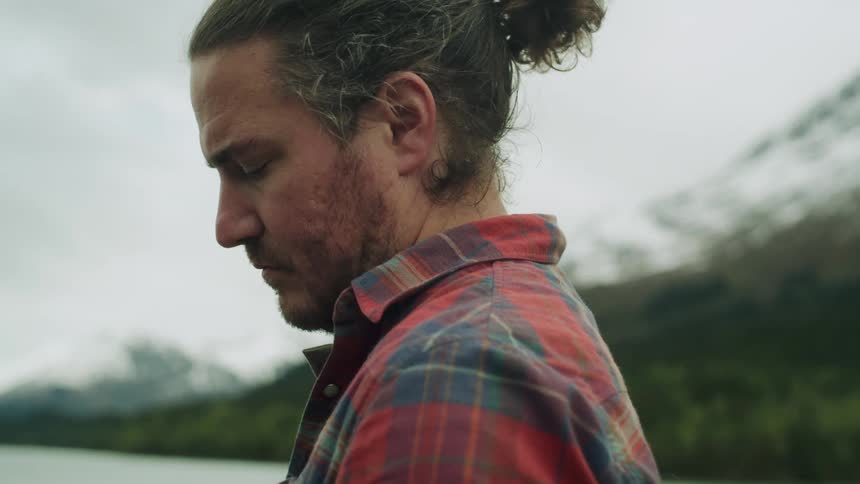 A man stands near the water with snow capped mountains and green trees on a cloudy day
