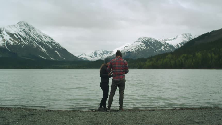 A man and a woman are standing together at the edge of a lake with snow covered mountains in the distance