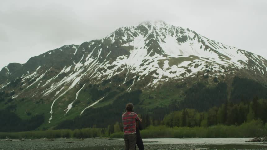 Man with long hair and plaid shirt holds woman watching high snowy mountain next to lake on a cloudy day