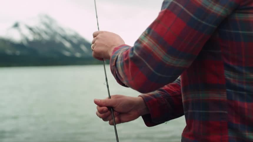 A man in front of a lake with a fishing rod with a mountain range the sky and clouds in the background