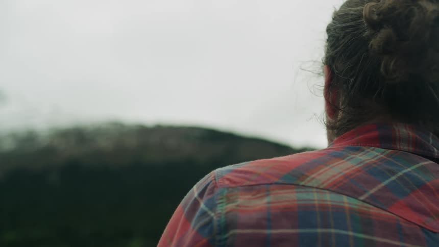 Man with long hair beard and red plaid shirt casts a fishing line into river