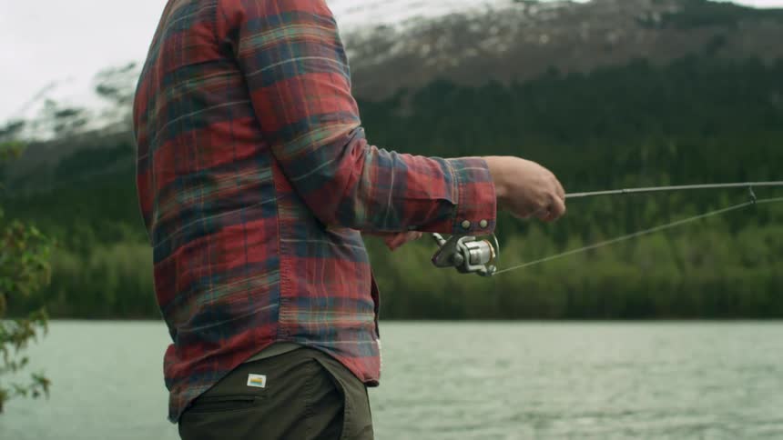 A man stands reeling in his fishing line near snow capped mountain and green trees