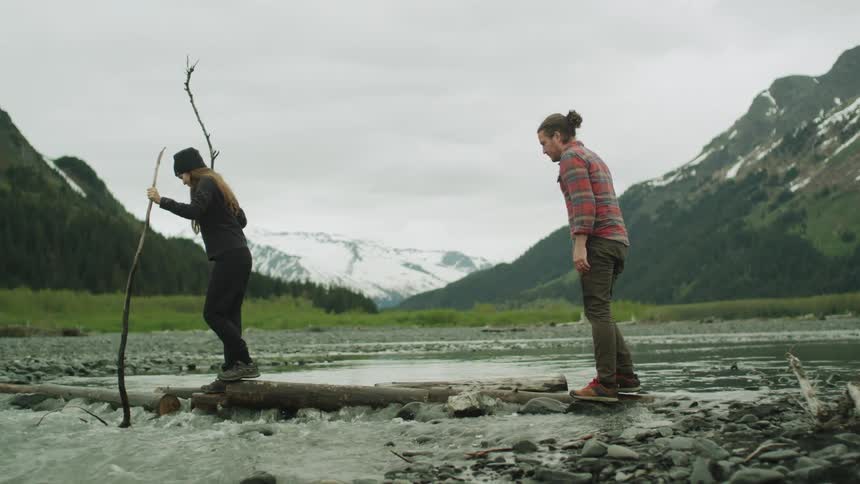 Man and woman carefully cross river on logs near slight slope with forest and mountains in background under an overcast sky