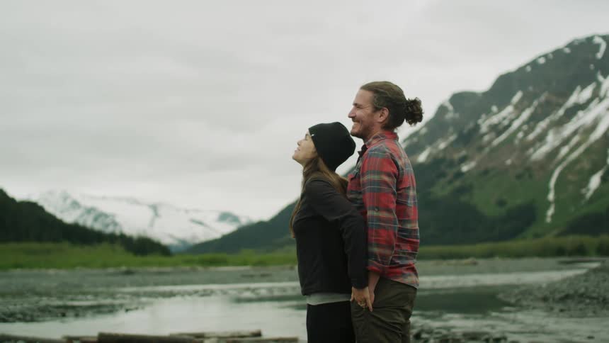 Couple stand against each other smiling with both of their hand in mans pocket looking at water and mountains