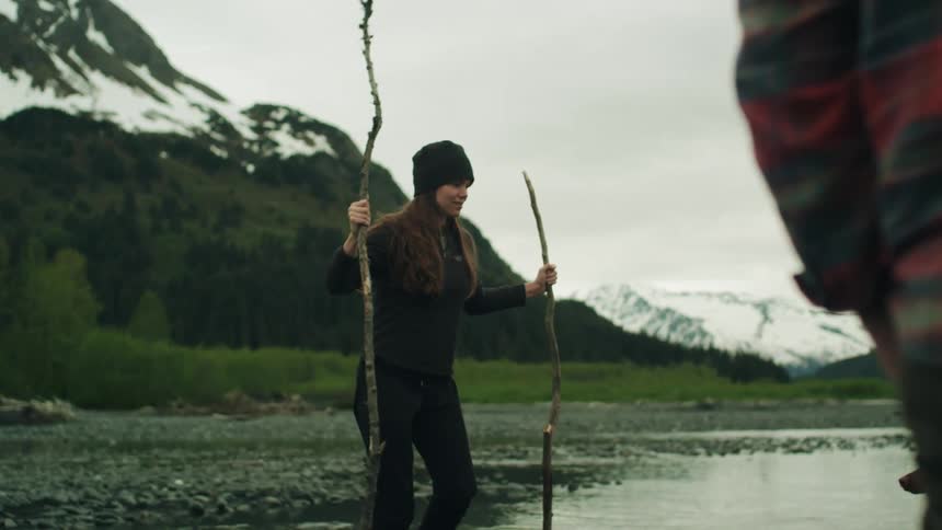 A woman uses two long branches to help her balance as she crosses a stream