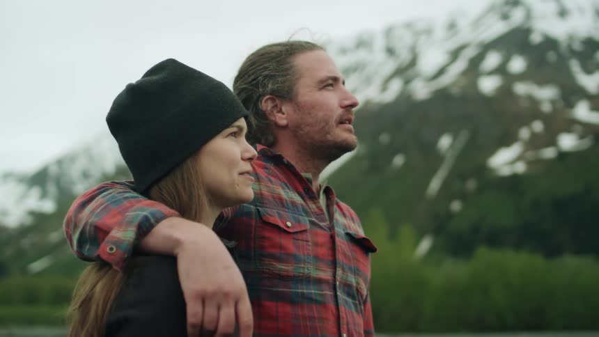 Woman with brown hair black beanie and sweater with man with long hair and red plaid shirt walk in the field with high mountains in the background
