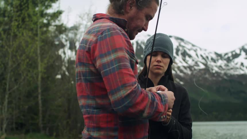 Woman wearing hat watches as man manipulates fishing line on fishing pole and water flows in background separating them from snowy mountains