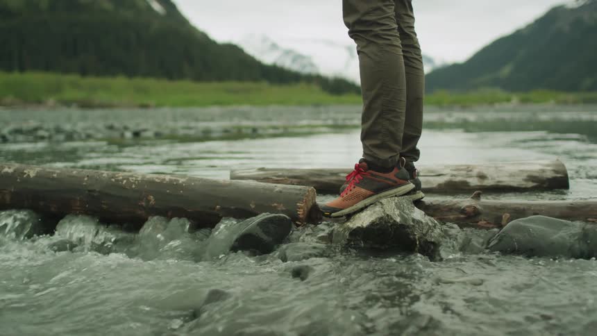 Sneakered feet stand on a rock near logs in a mountain stream as water flows by
