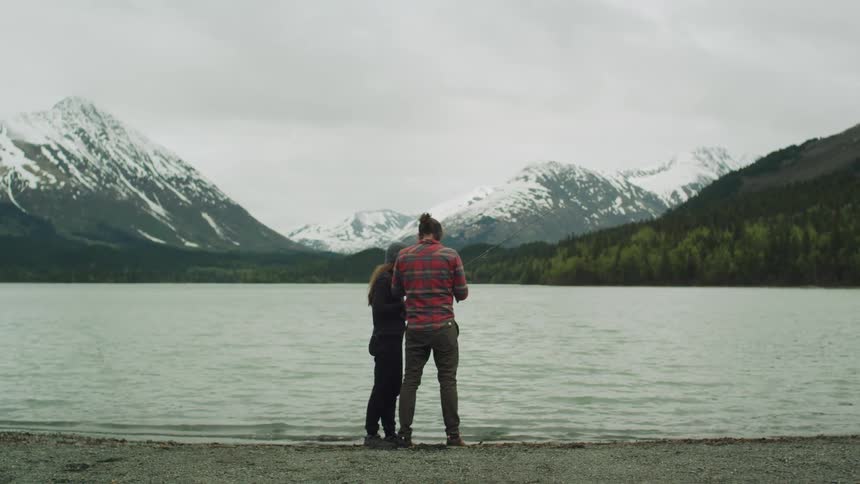 A man and woman stand at the edge of a lake near to snow covered mountains