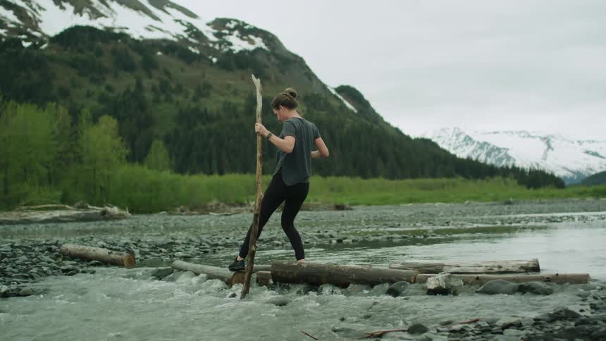 Woman carefully crosses river balancing on logs with aid of large stick with mountains in background