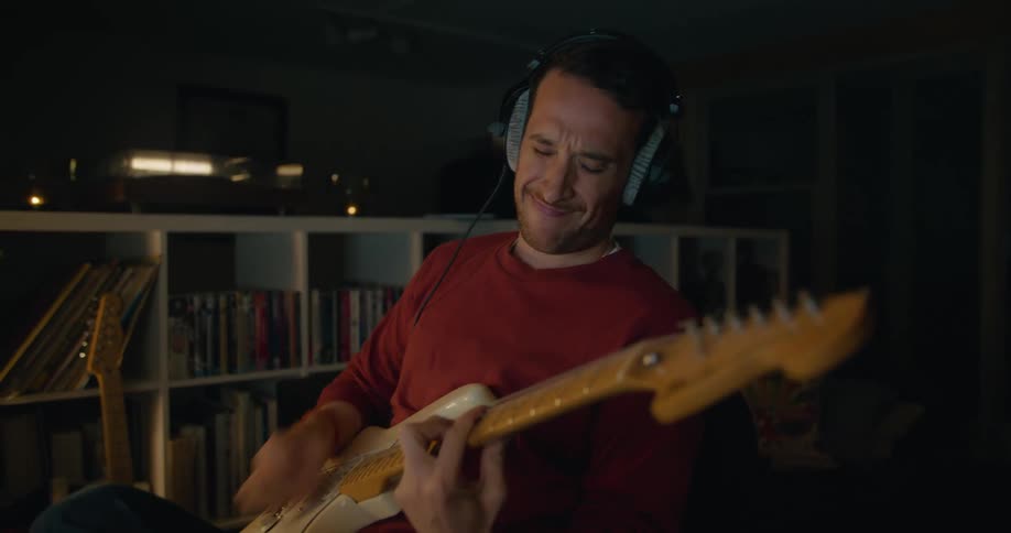A man plays guitar with headphones plugged in while sitting at home