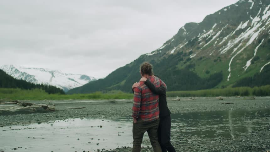 A woman smiles up at a man as she walks into his arms by the side of a mountain stream