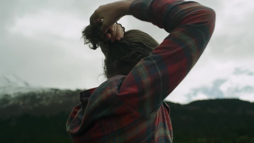 Man stands outdoors typing his hair up with mountains and forest in background