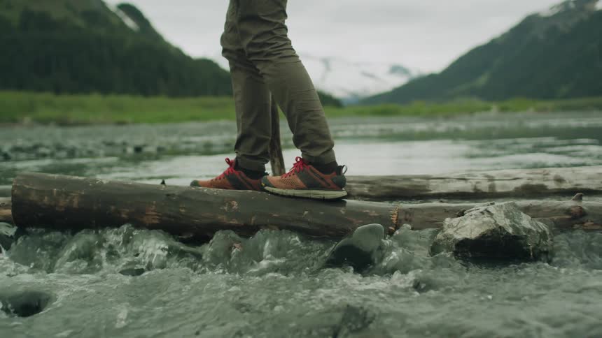 Man with green pants and orange black shoes walking across a river on tree trunk