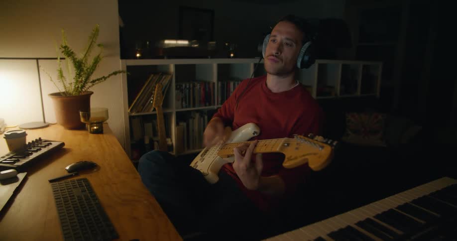 A male musician sits in a home studio while strumming on his electric guitar
