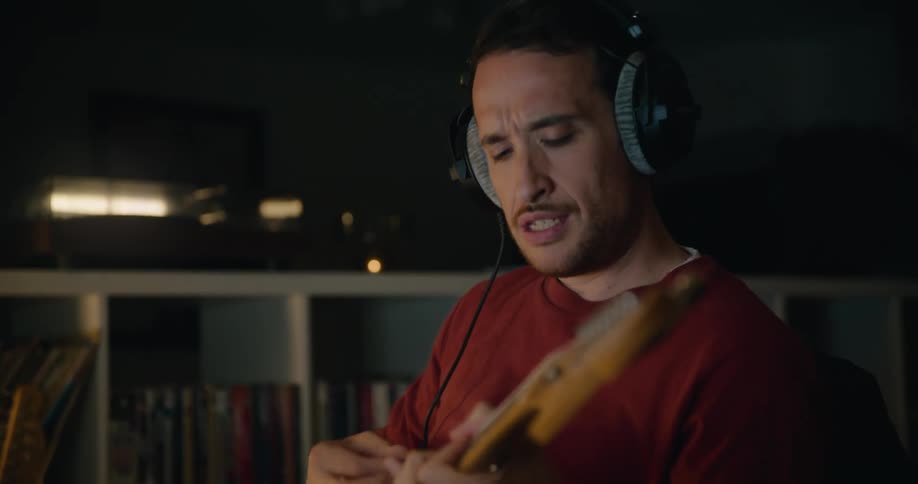 A man wearing headphones plays the guitar while sitting at a desk in a dimly lit room