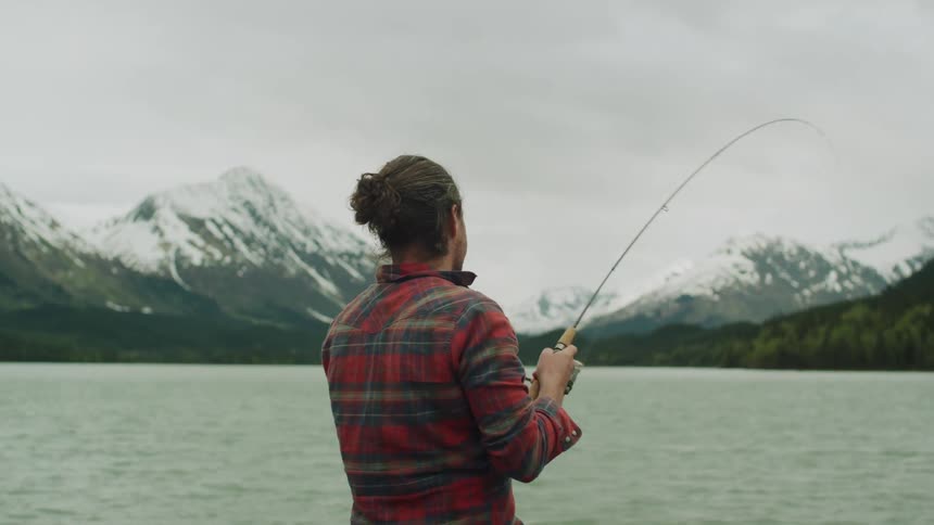 Man with red plaid shirt and long hair fish on lake next to snowy mountains