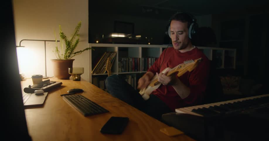 Man strums on guitar near piano keyboard and computer keyboard