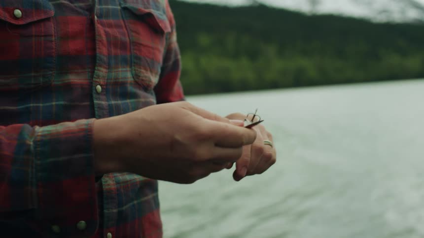 A man hooks bait on a fishing rod while at a lake near snow covered mountains