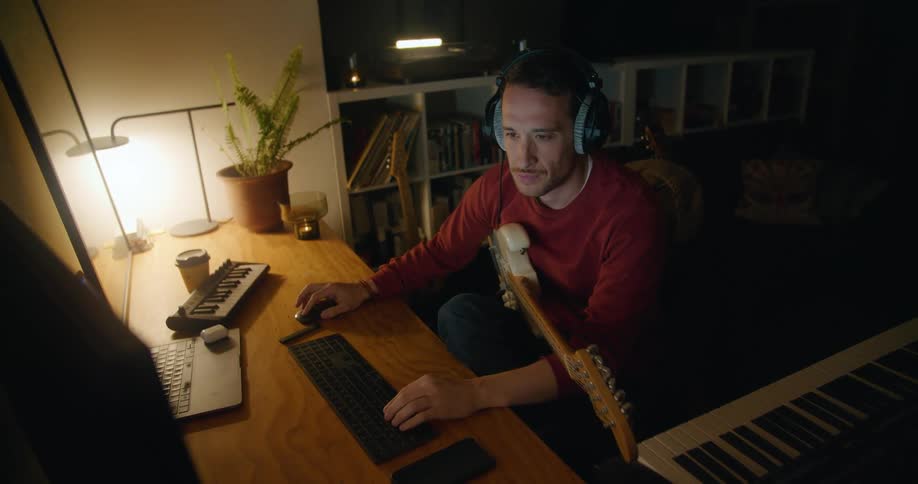 A man sits at a desk and plays his guitar and then uses his computer