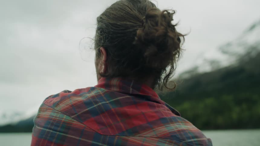 Man with long hair and red plaid shirt casts a fishing line into river