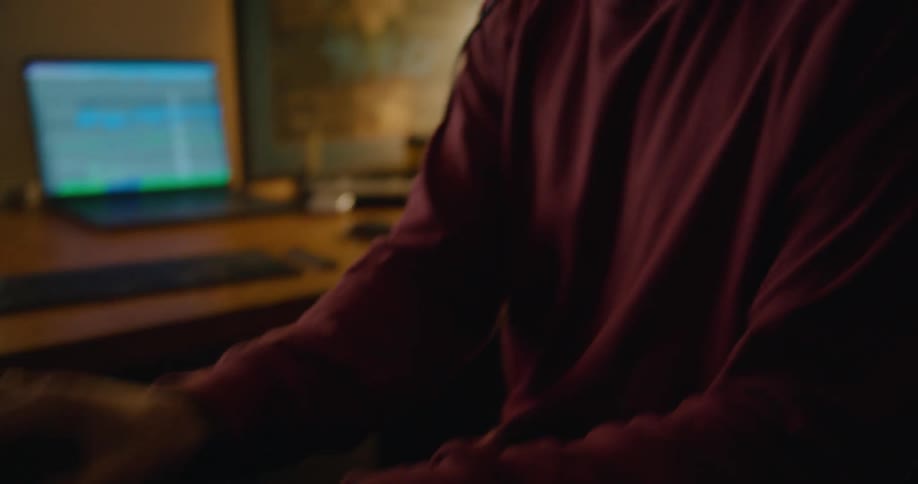 Musician wearing headphones bobs head as he play keyboard with laptop on nearby desk