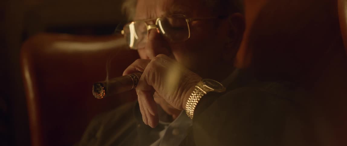 An older man wearing a gold watch puffs on a cigar