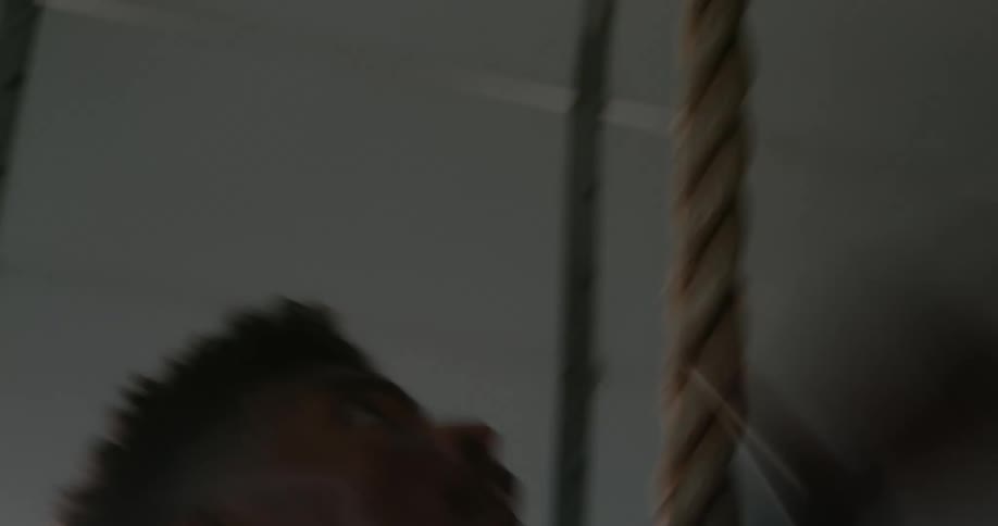 A man covered in a sweat climbs a rope in a gym for exercise