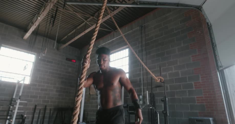 A muscular young man climbs down from a rope attached to the ceiling and prepares to start climbing it again