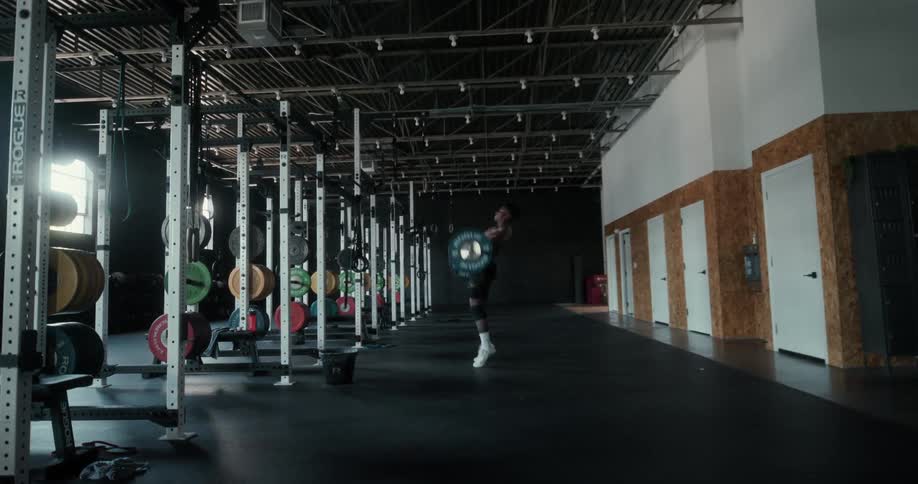 A young man in a large gym space is lifting large weights over his head